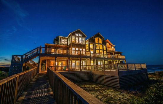 A twilight view highlighting the grandeur of the home, featuring multi-level wrap-around decks and a private wooden boardwalk leading directly to the beach.